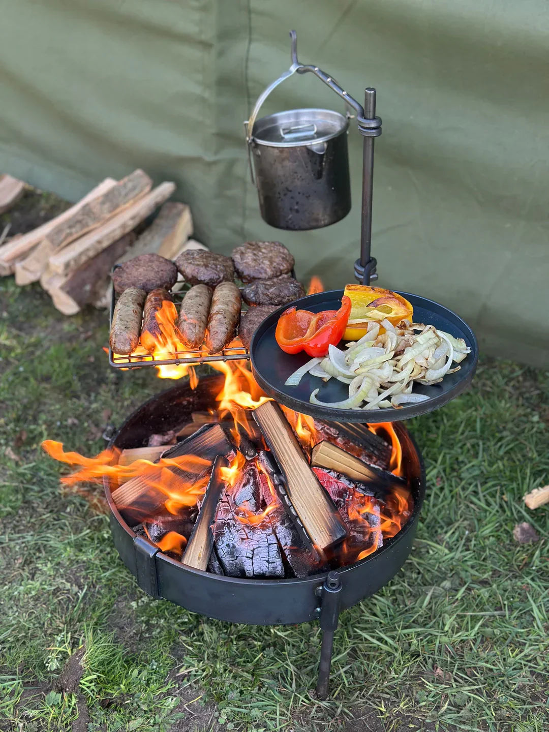Outdoor fire pit cooking setup with grilled meat, vegetables and a hanging pot