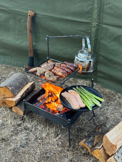 Outdoor cooking setup with meats, vegetables, and a kettle over a fire