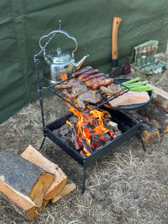 Outdoor cooking setup with meats on a grill over a fire, surrounded by camping gear.