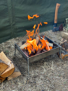Small fire pit with flames burning on a grassy area with logs and an axe in the background.