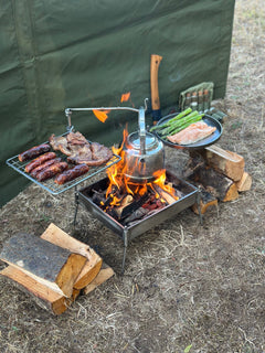 Outdoor cooking setup with meats, vegetables, and a teapot over a fire pit.